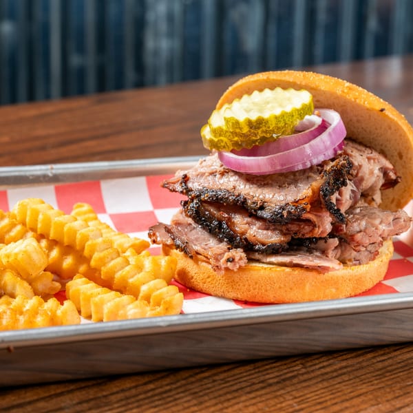Brisket sandwich with red onions and pickles, served with crinkle-cut fries on a metal tray.