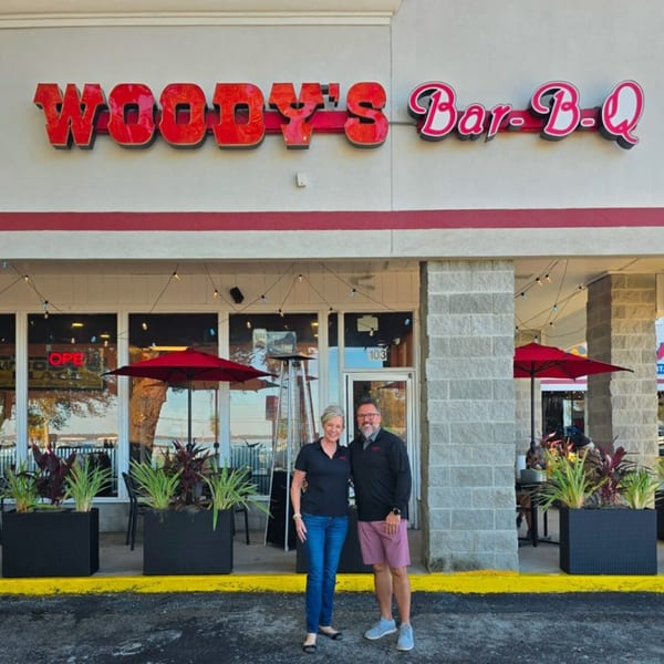 A man and woman stand smiling in front of Woody's Bar-B-Q restaurant entrance with outdoor seating.