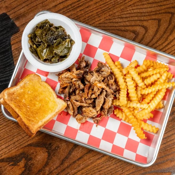 A metal tray with red and white checkered paper holds pulled pork, crinkle-cut fries, collard greens, and toast.