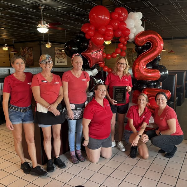 Seven Woody's Bar-B-Q staff members in red shirts pose with a 25th-anniversary plaque and balloons.