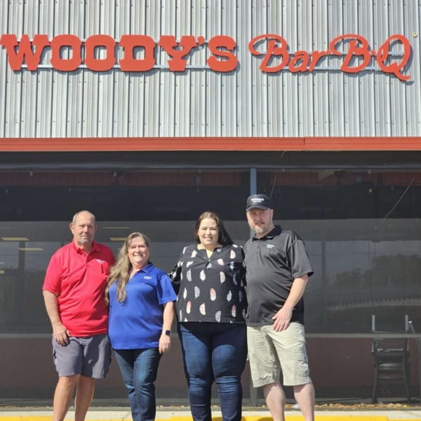 Four people stand smiling in front of a Woody's Bar-B-Q restaurant building with a red sign.