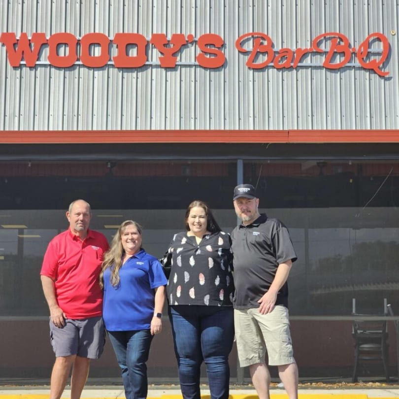 Four people stand smiling in front of a Woody's Bar-B-Q restaurant building with a red sign.