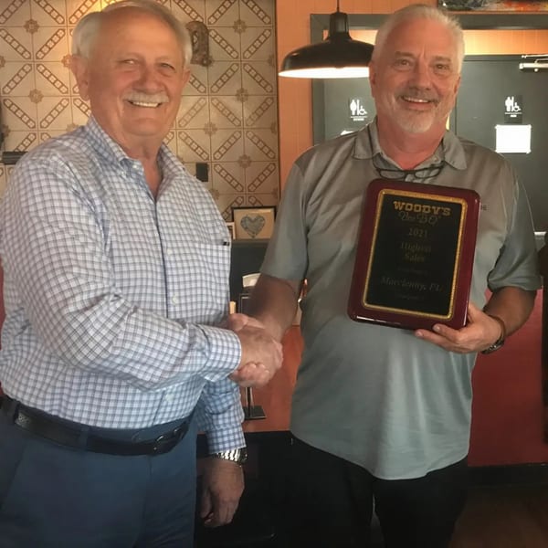 Two men shaking hands, one holding a "Woody's BBQ Highest Sales" award plaque.