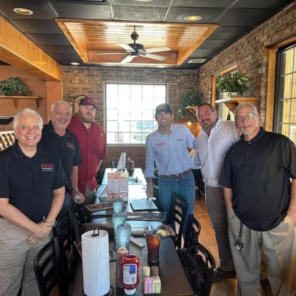 Six men smiling and posing for a photo in a restaurant dining area with brick walls.
