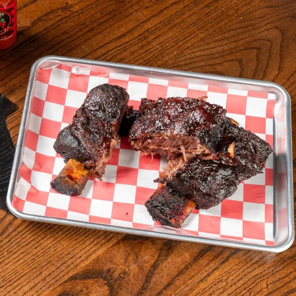 Two large, dark, glazed short ribs on a metal tray with red and white checkered paper.